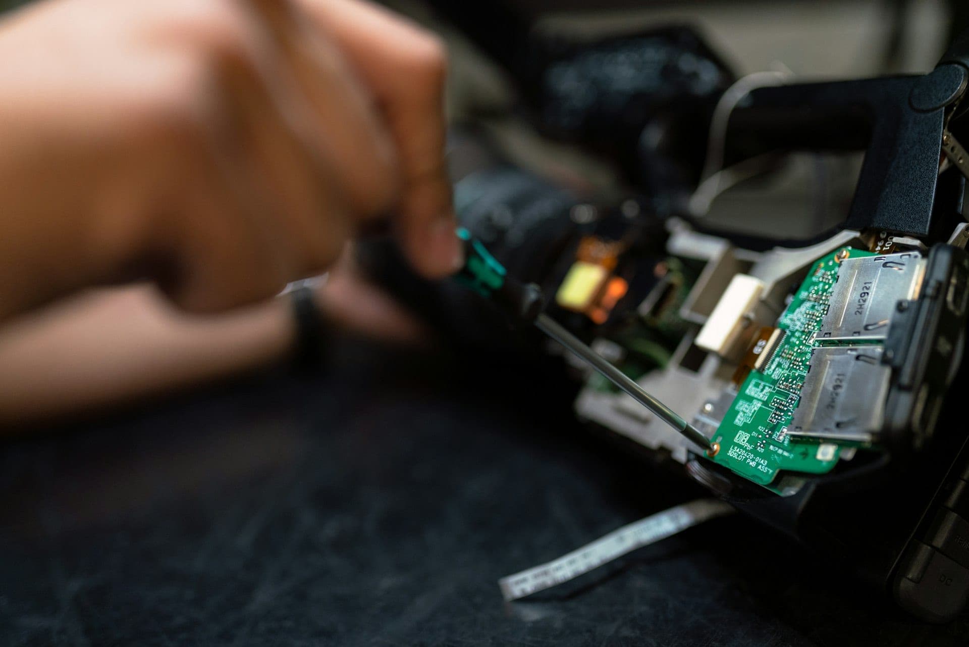 A technician's hands working on the opened circuit board of an electronic device at a repair bench.